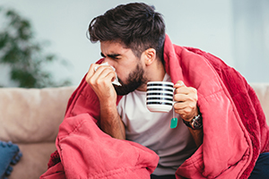 Man blowing his nose while holding a cup of warm coffee.