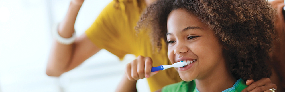 Young girl brushing teeth with mother standing behind her.