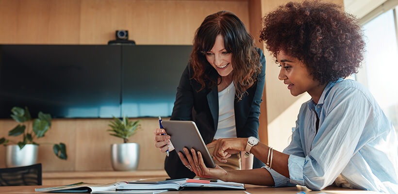 Two women are working together on a tablet in a conference room.