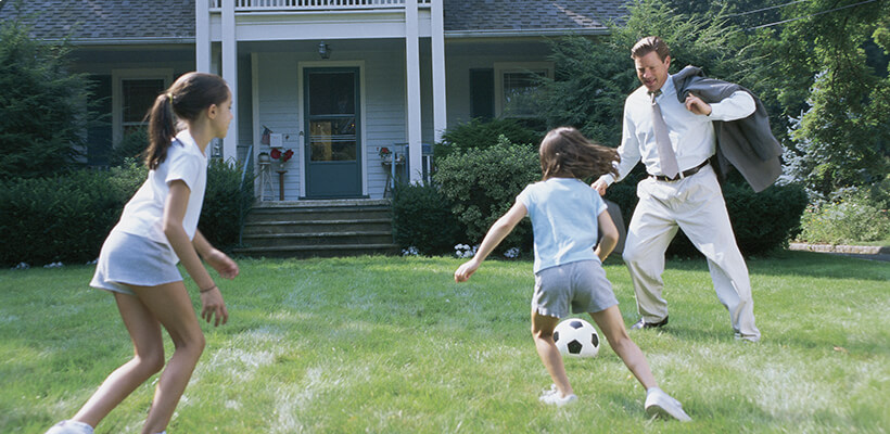 A dad plays soccer with his two daughters in the front yard of their house after he gets home from work.