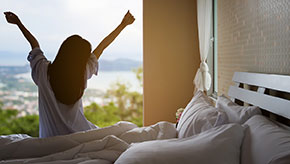 A woman stretches in bed as she wakes up with the sun rising through her window.
