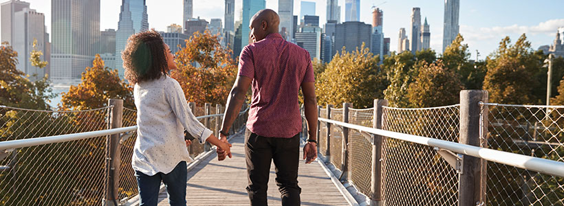 A couple in their early 30s practice healthy lifestyle habits by going for a walk over a pedestrian bridge toward a city in front of them.