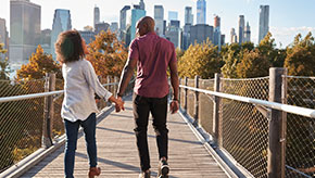 A couple in their early 30s practice healthy lifestyle habits by going for a walk over a pedestrian bridge toward a city in front of them.