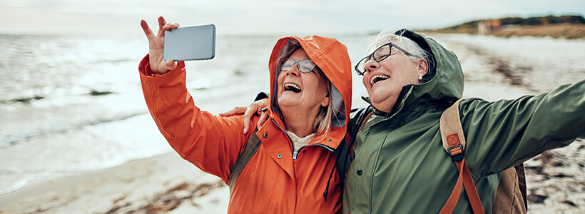 Two retired women practice mindfulness by living in the moment, spreading their arms in joy together and take a selfie on the beach.