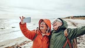 Two retired women practice mindfulness by living in the moment, spreading their arms in joy together and take a selfie on the beach.