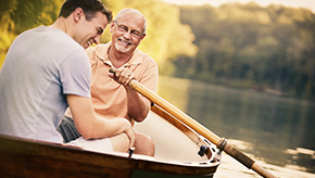 A grandfather shares a laugh with his teenage grandson while rowing a small boat on a lake on a sunny, spring day.