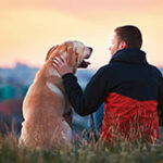 A man petting his dog while sitting in the grass overlooking a scenic view.