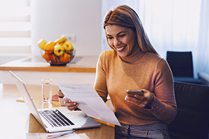 Cute caucasian smiling blonde woman in sweater holding bills in one hand and in other smart phone. On table are laptop and bills. Apartment interior.