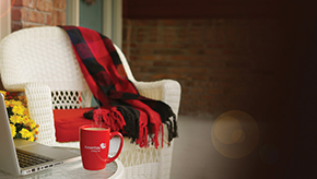 Empty wicker chair sits on a porch with blanket draped over the back near a table holding a laptop and coffee cup.