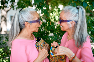 Senior women twins in colorful clothes with extraordinary handbags in city, talking and smiling.