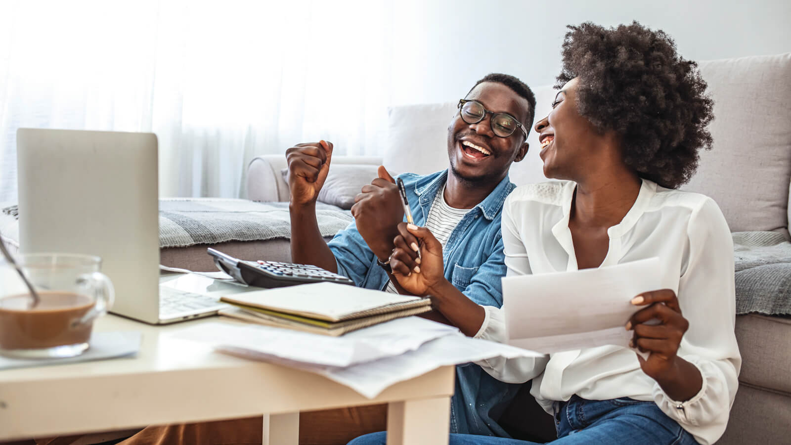 A young couple relax as they review their personal finances by checking paperwork and a financial checklist on their laptop.