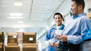 Two employees take a coffee break in a warehouse. They’re relaxed because their employer recently enrolled in a PEP to help with employees’ retirement planning.