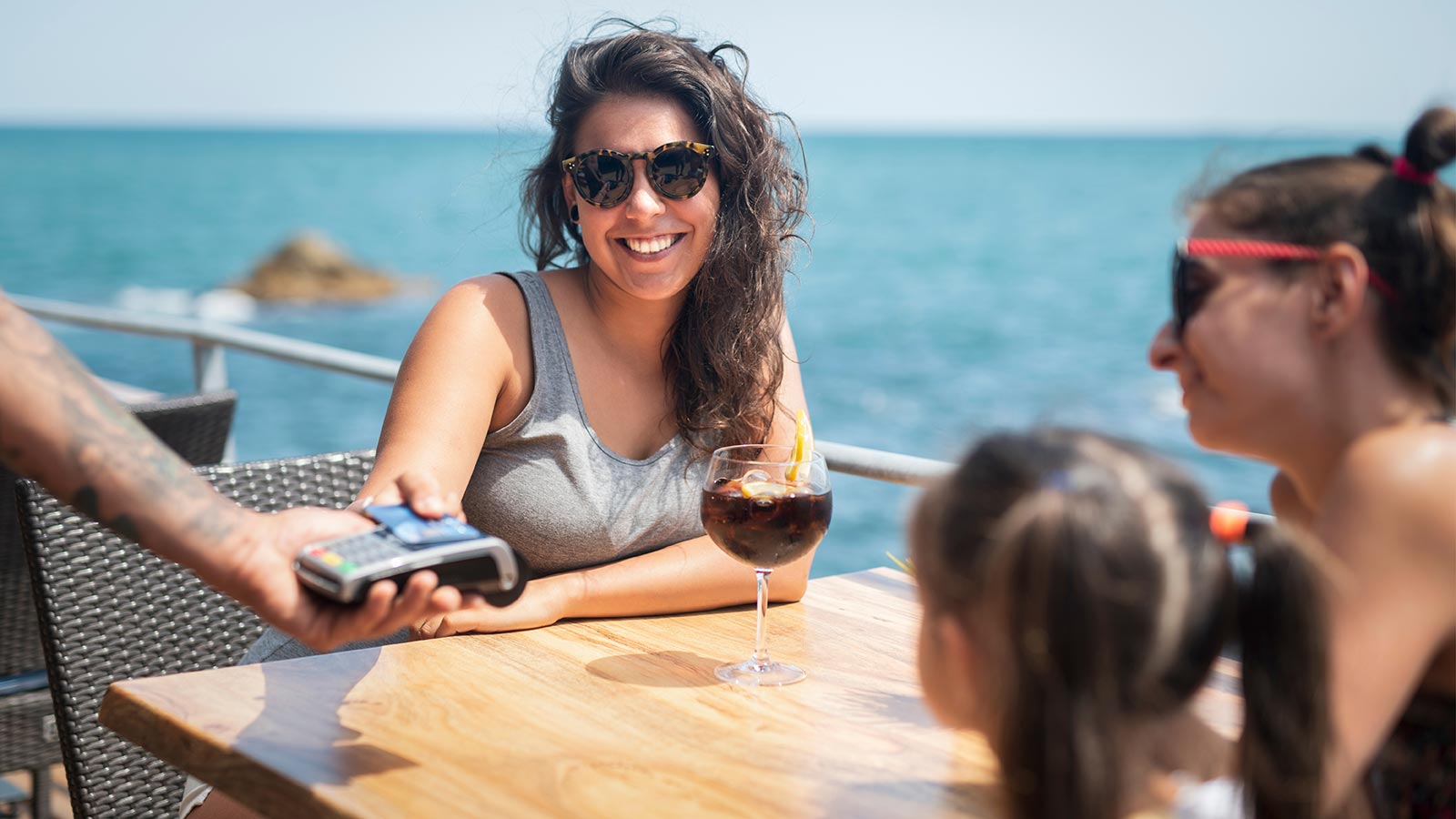 A woman sits in the sun enjoying a summer vacation and pays for a drink with her credit card to keep her money safe while she travels.