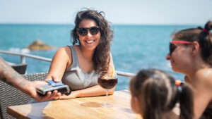 A woman sits in the sun enjoying a summer vacation and pays for a drink with her credit card to keep her money safe while she travels.