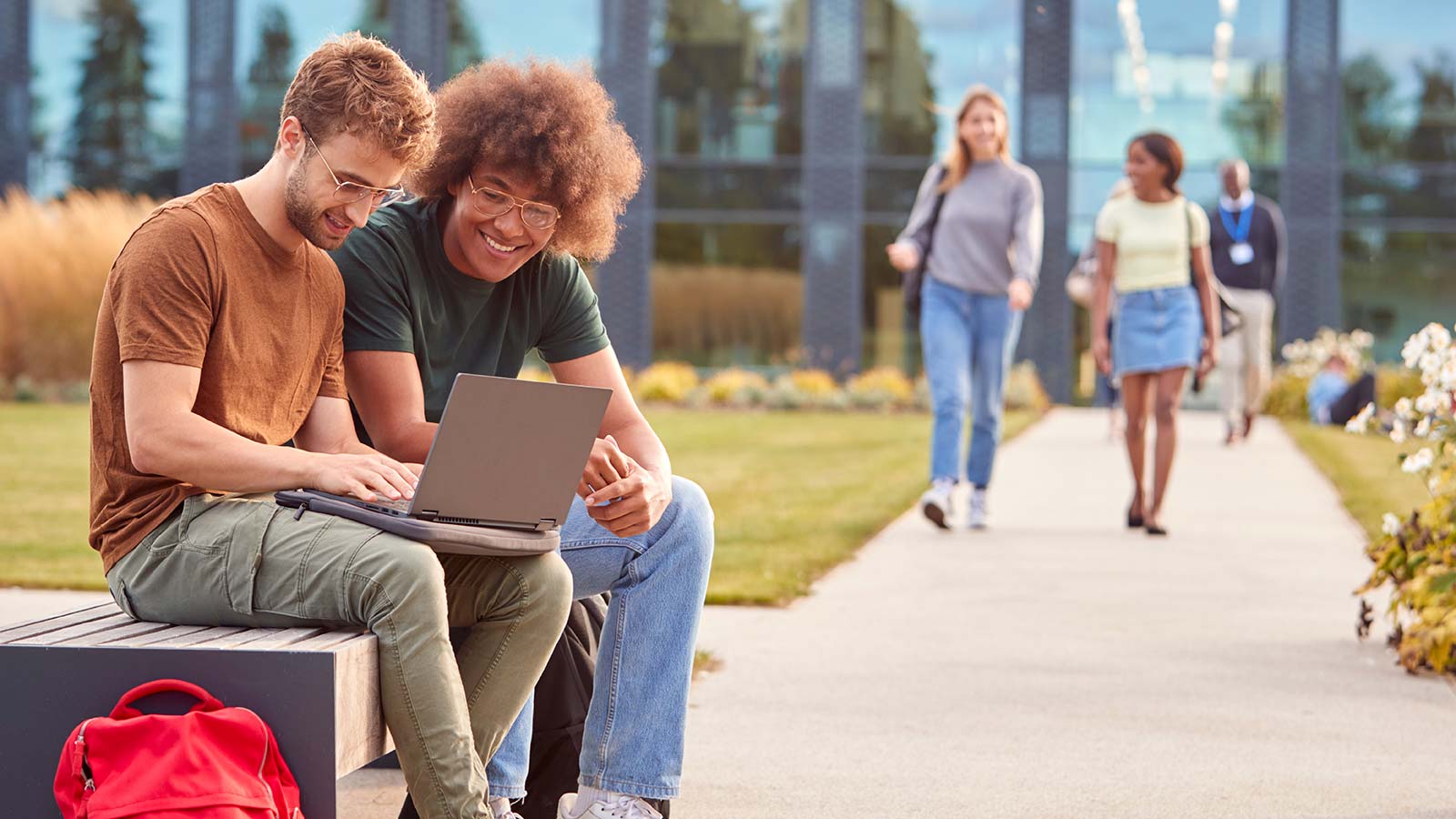Two young men sit on a bench outside on their college campus and review a project on a laptop before walking to class.