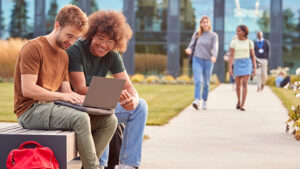 Two young men sit on a bench outside on their college campus and review a project on a laptop before walking to class.