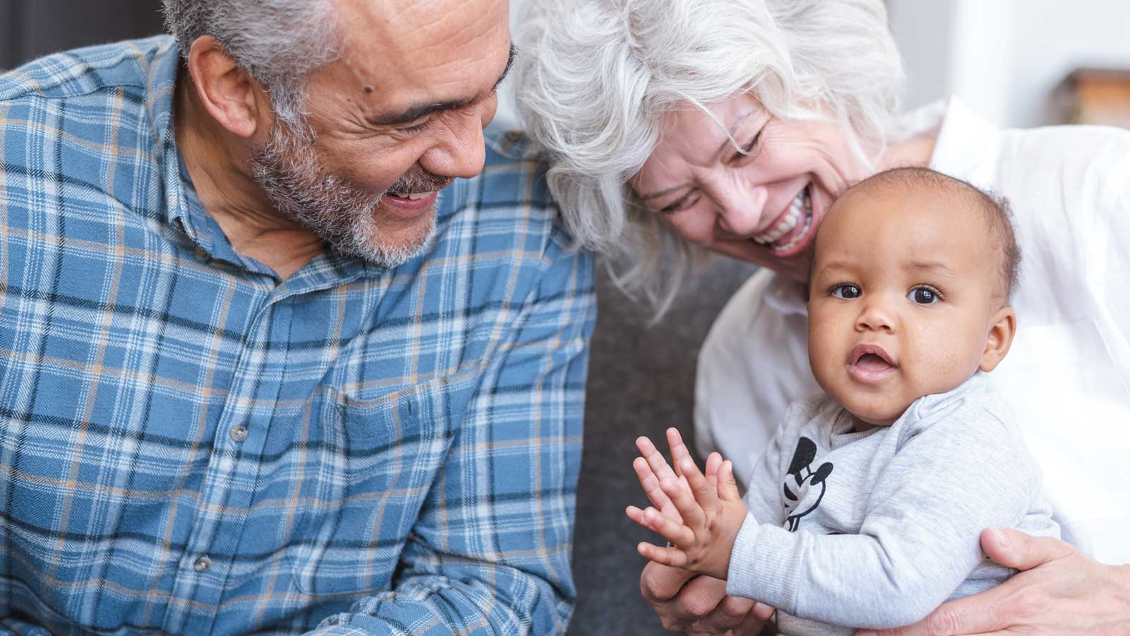 A baby boy sits on his grandma’s lap clapping his hands. Grandma and grandpa smile and watch him happily.