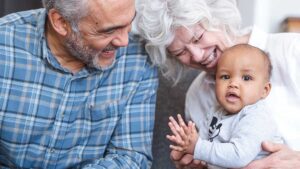 A baby boy sits on his grandma’s lap clapping his hands. Grandma and grandpa smile and watch him happily.