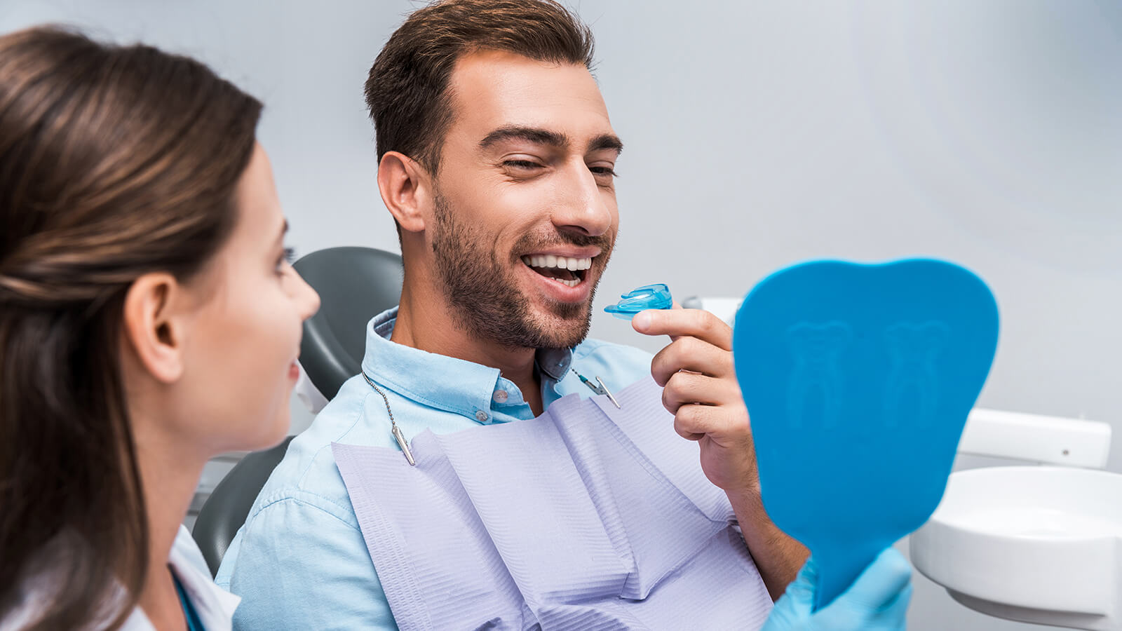 Male sitting in dental chair smiling into a hand held mirror.