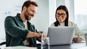 Male and female sitting at a table, smiling, and viewing a laptop.