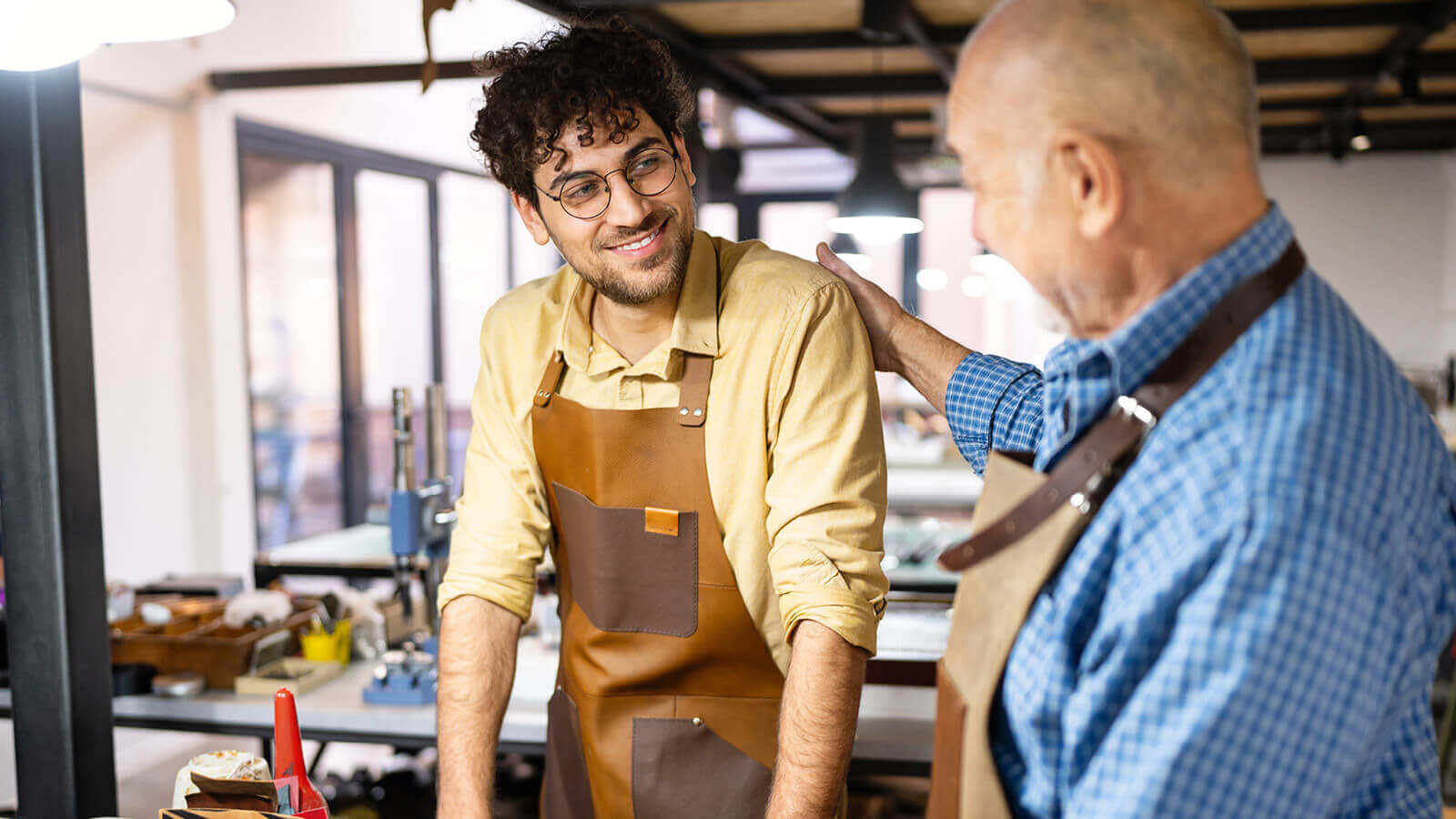 A father proudly looks at his son working alongside him in the workshop of their family business.