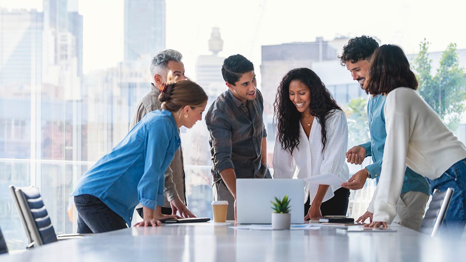 A group of colleagues in an office gathered around a table looking at a laptop.