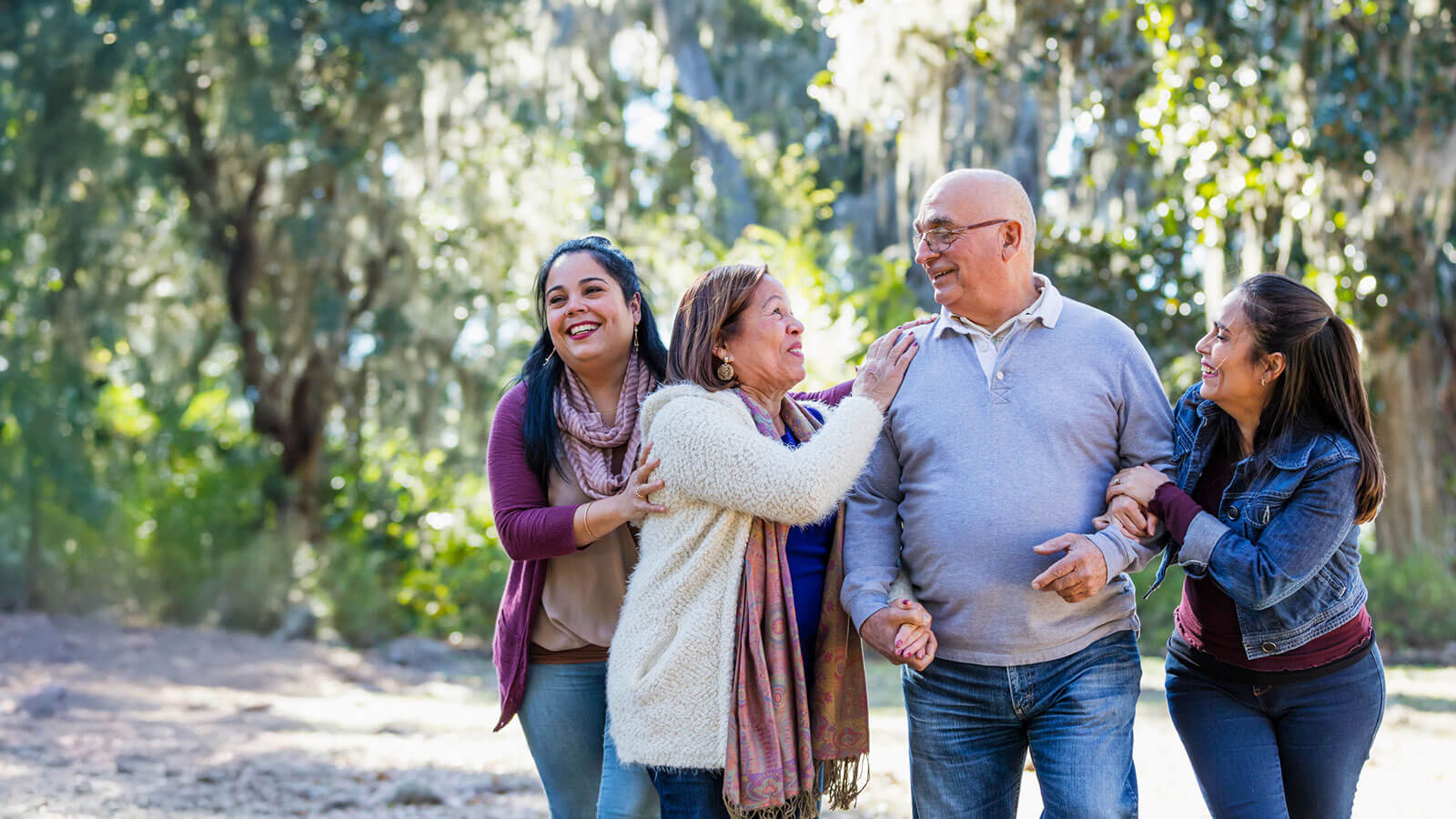 A family of four, two parents and their two adult daughters, are taking a walk on a sunny, fall day through a wooded park, laughing and having a fun afternoon together. A family of four, two parents and their two adult daughters, are taking a walk on a sunny, fall day through a wooded park, laughing and having a fun afternoon together.