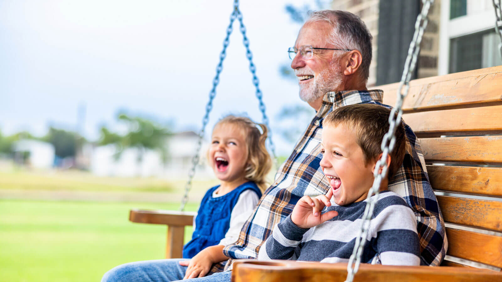 A retired grandfather sits on a front porch swing with his two young grandchildren on either side of him, and all three of them are smiling. A retired grandfather sits on a front porch swing with his two young grandchildren on either side of him, and all three of them are smiling.