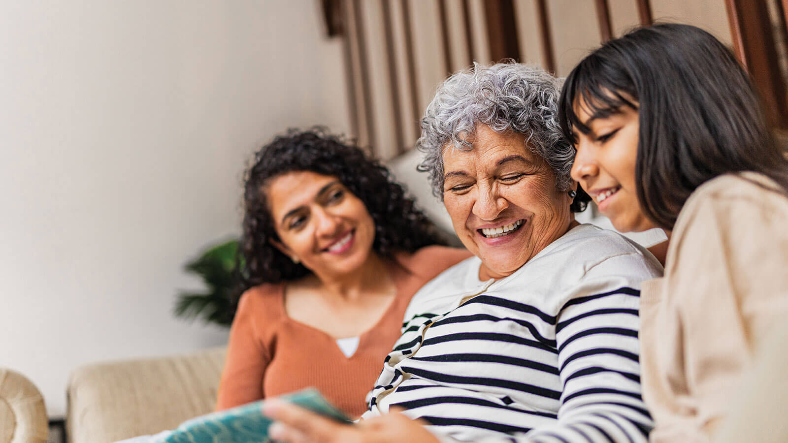 A grandmother sits on the couch with her adult daughter and young granddaughter looking at a family photo album.