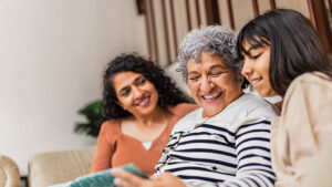 A grandmother sits on the couch with her adult daughter and young granddaughter looking at a family photo album.