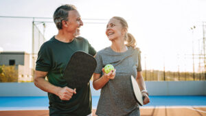 A husband and wife in their late 60s walk off a pickleball court enjoying their retirement hobby.