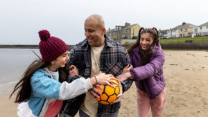 A dad in his 40s plays soccer on the beach with his teen and pre-teen daughters.