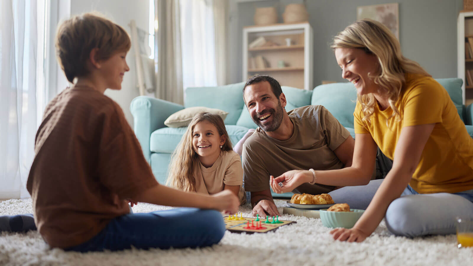 A young family of four are all smiling happily playing a board game casually on the floor.