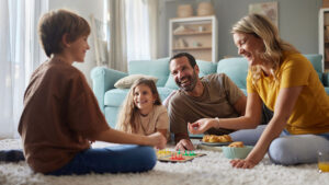 A young family of four are all smiling happily playing a board game casually on the floor.