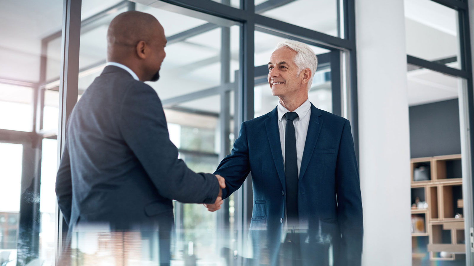 Two business professionals in suits shaking hands in a modern glass‑walled office.