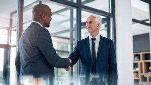 Two business professionals in suits shaking hands in a modern glass‑walled office.