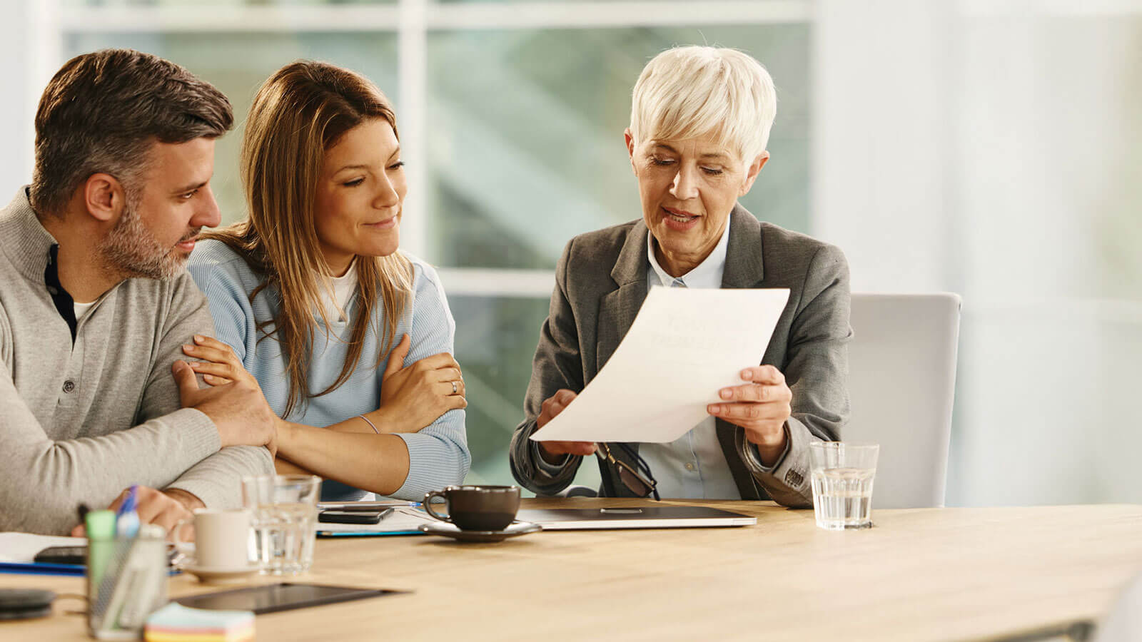 Female financial professional with short gray hair reviews documents with a couple during a meeting in a modern office.