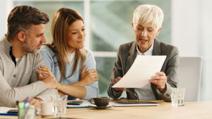 Female financial professional with short gray hair reviews documents with a couple during a meeting in a modern office.