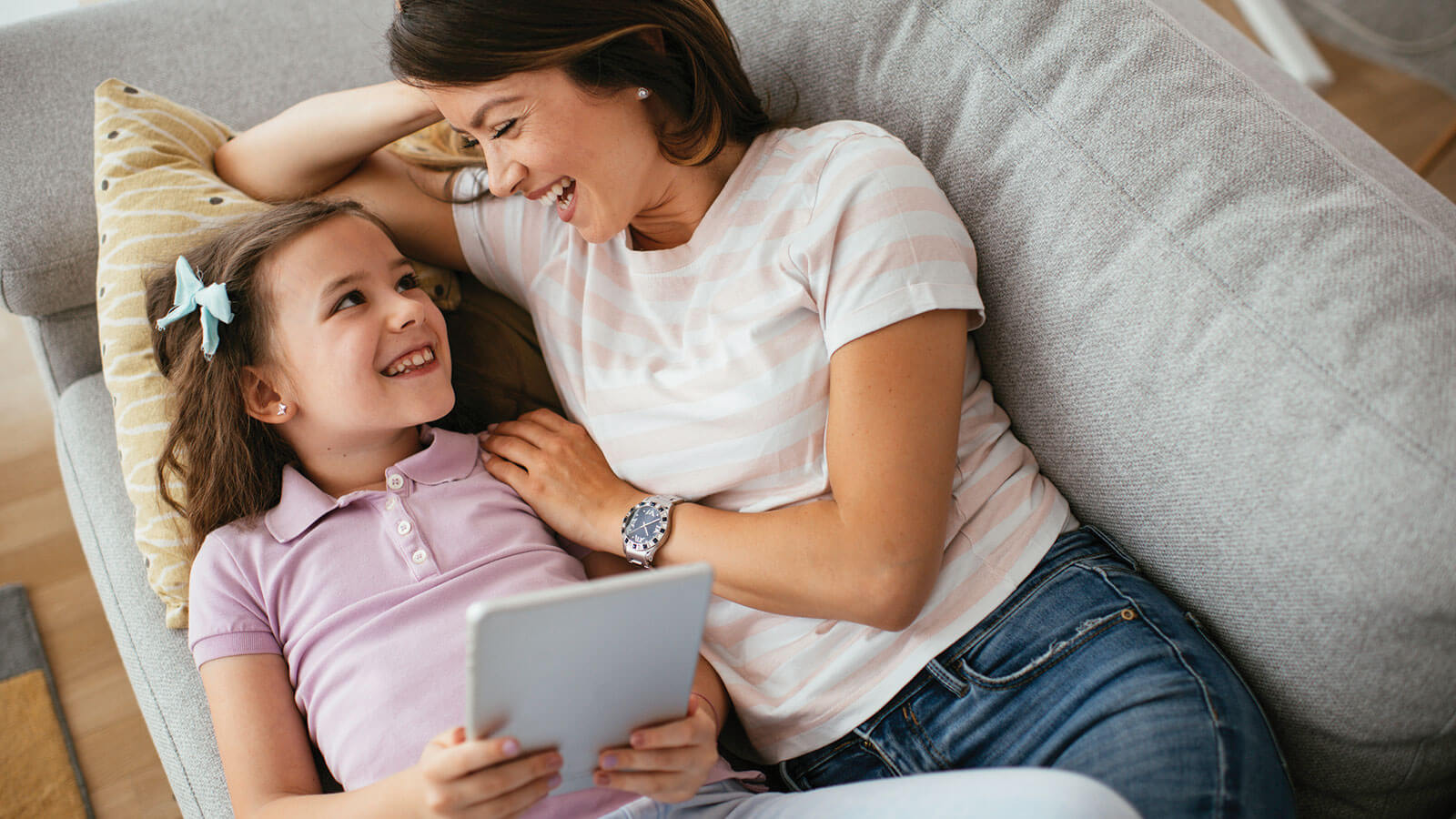 A mom and her young daughter snuggle on the couch laughing at something on the daughter’s tablet.