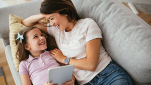 A mom and her young daughter snuggle on the couch laughing at something on the daughter’s tablet.