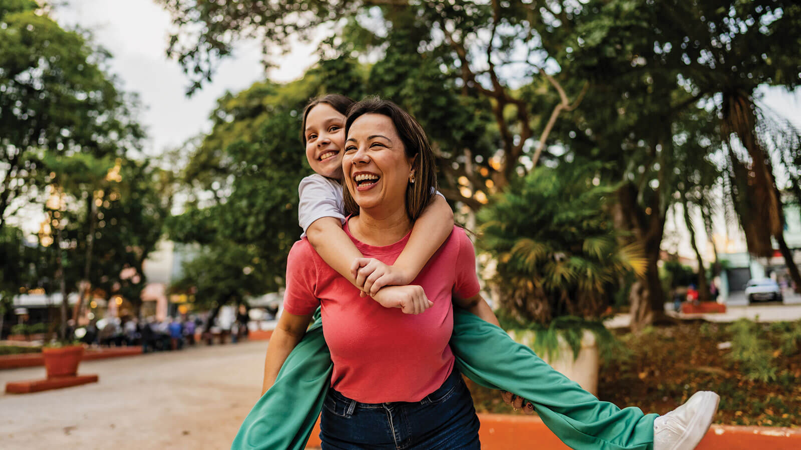 A mom laughs giving her daughter a piggyback ride while walking through a park, illustrating family responsibilities and long-term financial goals.