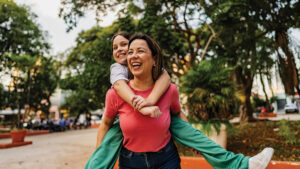 A mom laughs giving her daughter a piggyback ride while walking through a park, illustrating family responsibilities and long-term financial goals.