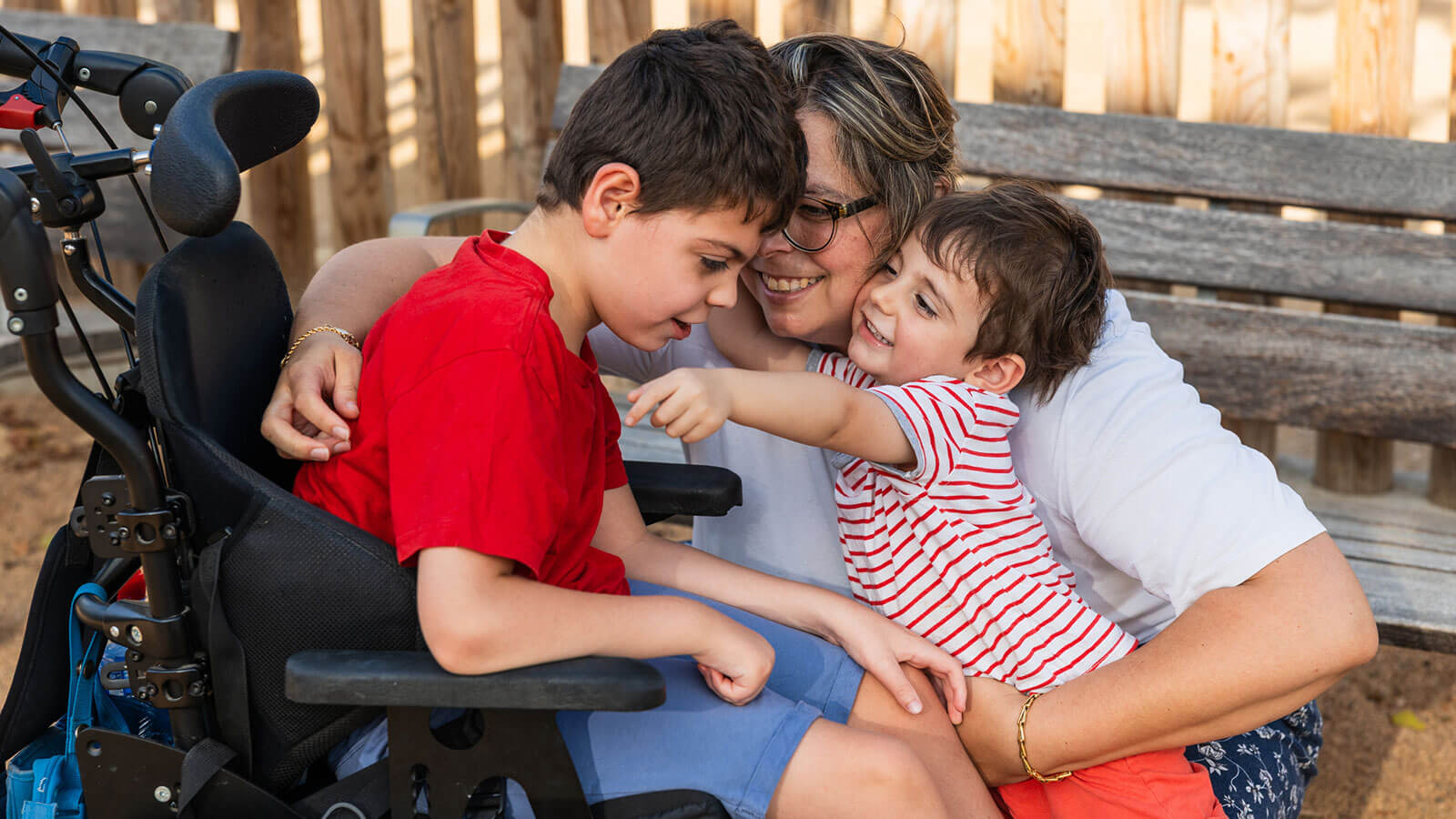A mom plays with and hugs her two sons outside at a park. One child has special needs using a wheelchair, reflecting the importance of planning for the future care of a loved one with special needs.
