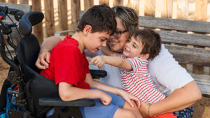 A mom plays with and hugs her two sons outside at a park. One child has special needs using a wheelchair, reflecting the importance of planning for the future care of a loved one with special needs.