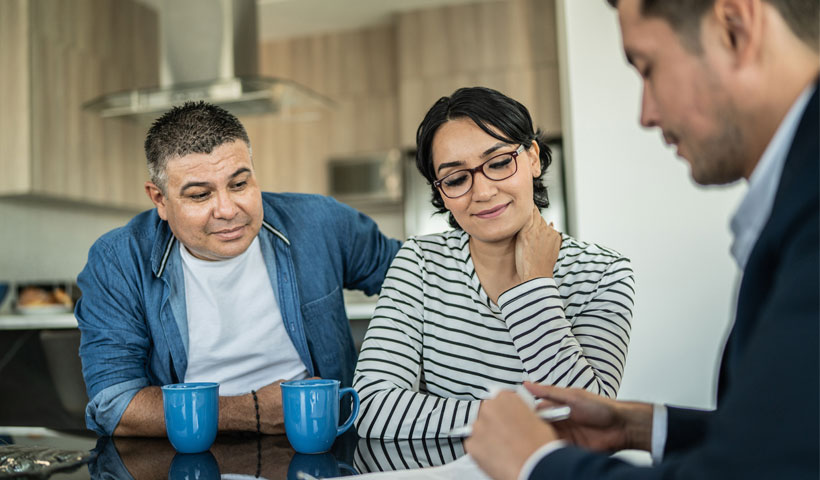 Three people sit around a kitchen table with blue mugs as one person gestures toward papers during a face-to-face conversation.