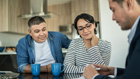 Three people sit around a kitchen table with blue mugs as one person gestures toward papers during a face-to-face conversation.